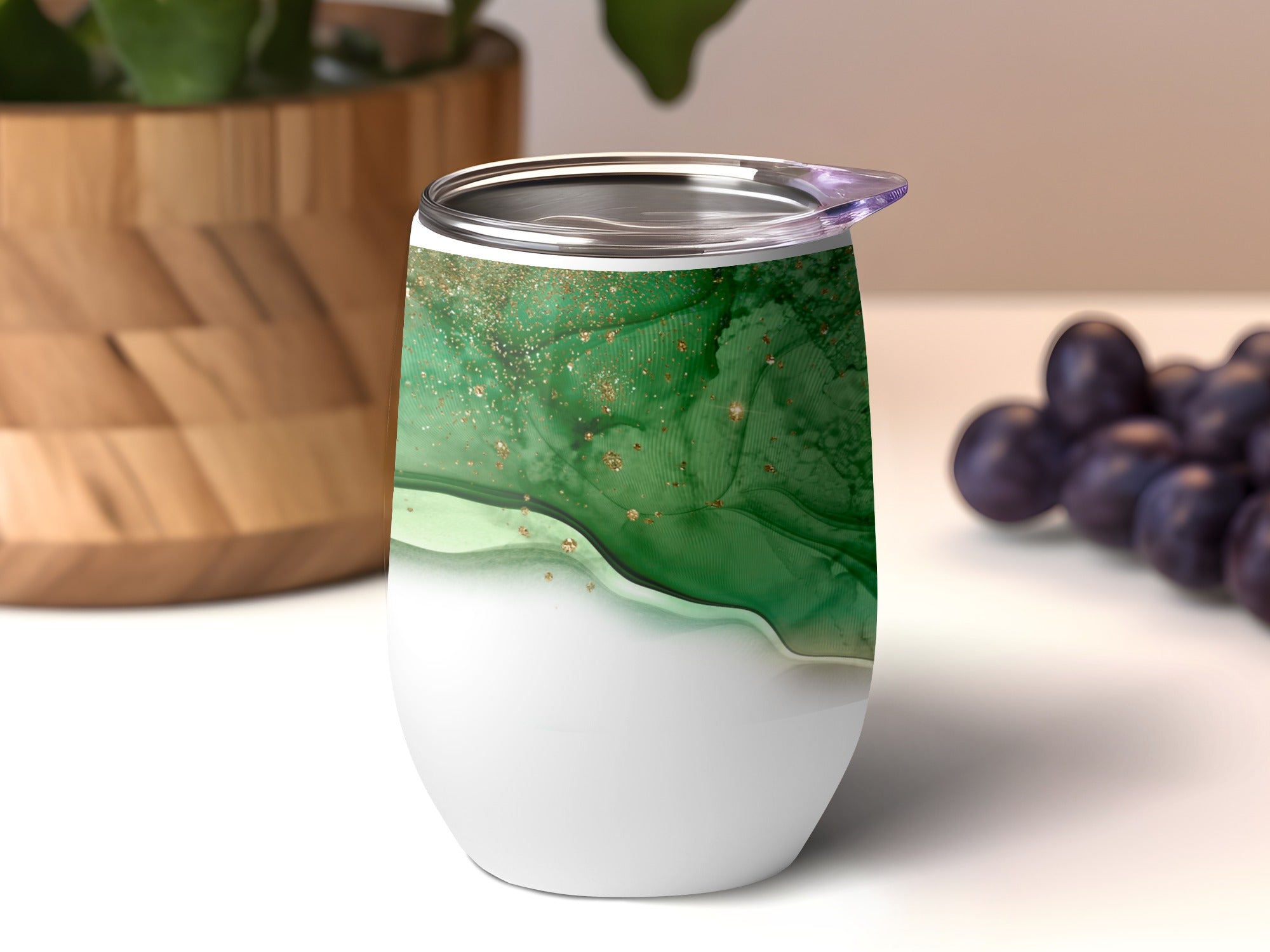 White tumbler with green and gold abstract design, blurry grapes and plant in background.