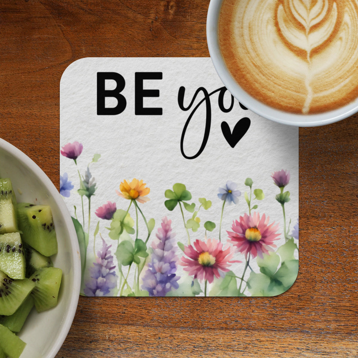 White coaster with watercolor flowers and "BE you" text beside coffee and kiwi.
