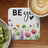 White coaster with watercolor flowers and "BE you" text beside coffee and kiwi.