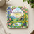 Square coaster with a landscape, water, flowers, and "God is our refuge and strength" Psalm 46:1.