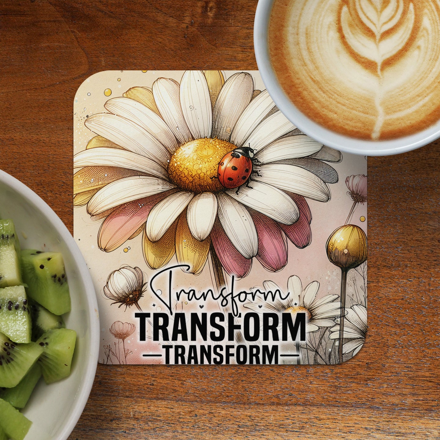Daisy with ladybug and text "TRANSFORM" on a coaster next to coffee and kiwi.