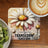 Daisy with ladybug and text "TRANSFORM" on a coaster next to coffee and kiwi.