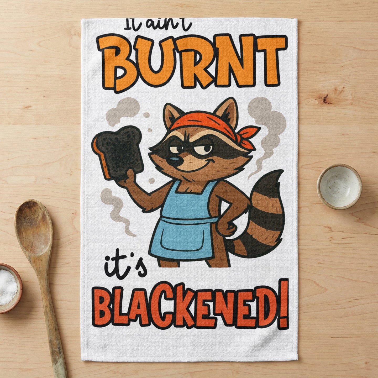 Raccoon chef in apron holding burnt toast with text "It ain't burnt, it's blackened!".