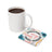 White mug with coffee next to a round coaster with floral design and text.