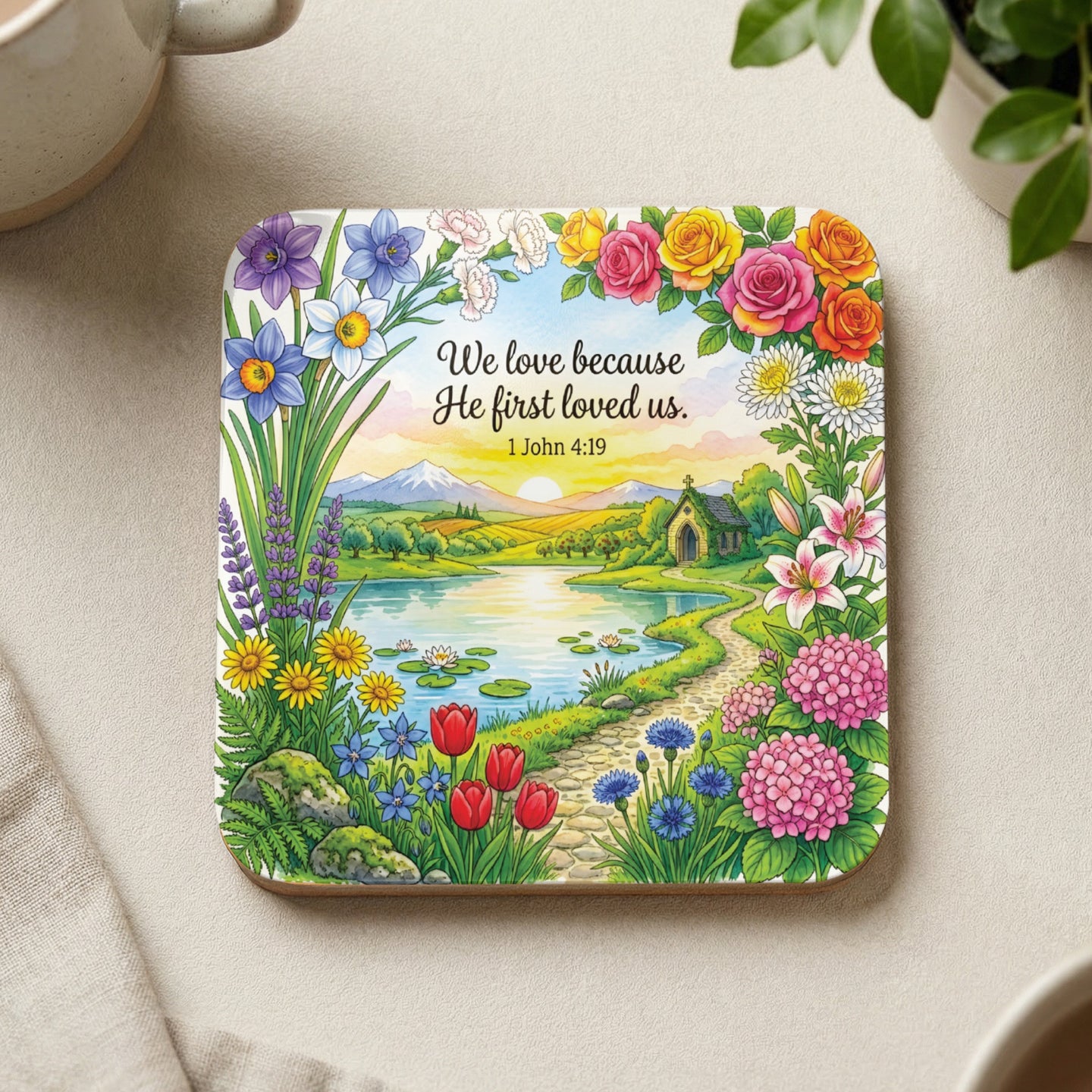 Square coaster with a scenic landscape, flowers, and text "We love because He first loved us. 1 John 4:19".
