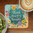 Coaster with "Think Happy Thoughts" in colorful floral design next to kiwi and coffee.