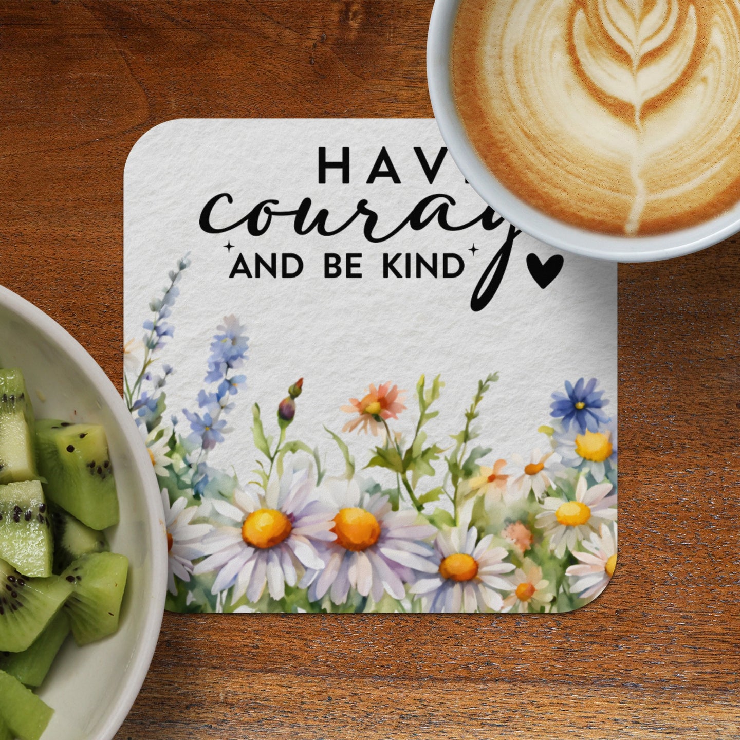 Coaster with floral design and "Have courage and be kind" text next to coffee and kiwi.