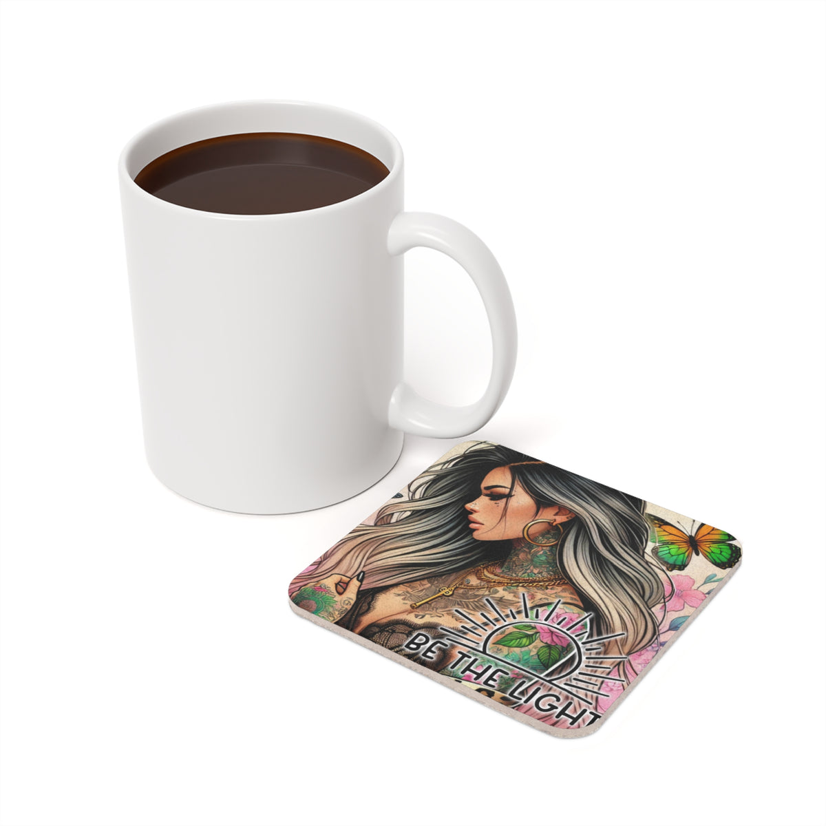 White mug next to square coaster with tattoo art and "BE THE LIGHT" text.