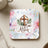 Square coaster with "He is Risen" text, a lamb, and a cross surrounded by flowers.