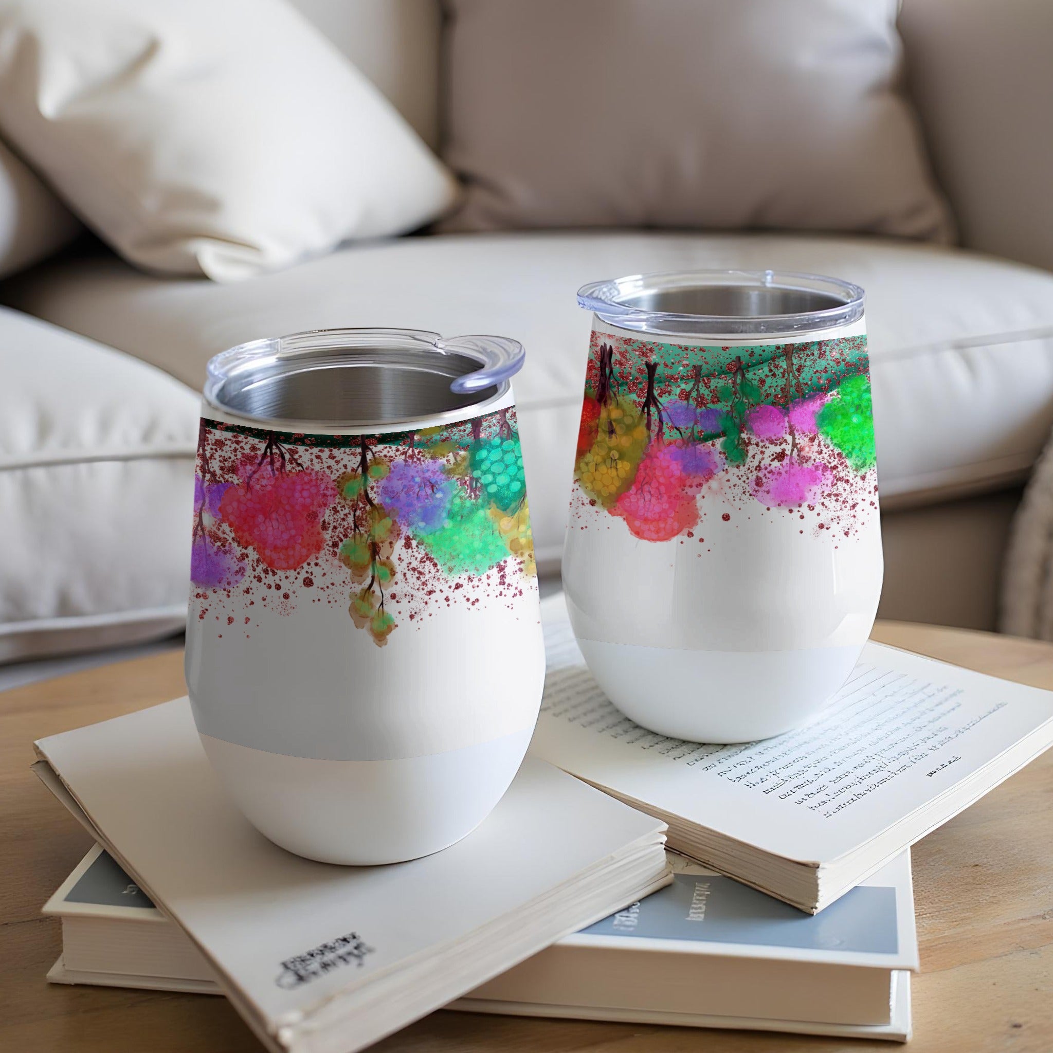 Two white tumblers with colorful abstract paint splatter design sit on books.