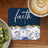 Dark blue coaster with "faith" and floral pattern next to fruit and coffee.