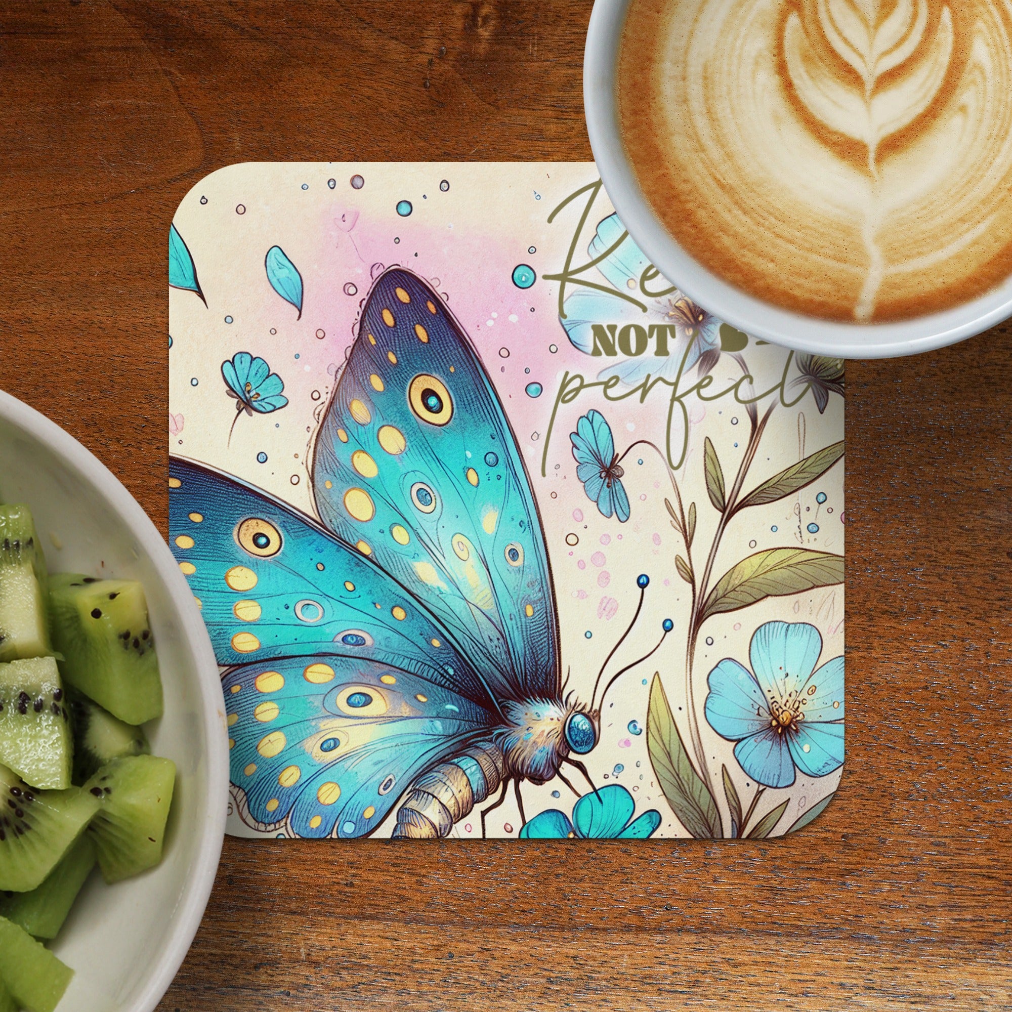 Blue butterfly with floral design on coaster, next to kiwi and coffee.