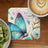 Blue butterfly with floral design on coaster, next to kiwi and coffee.