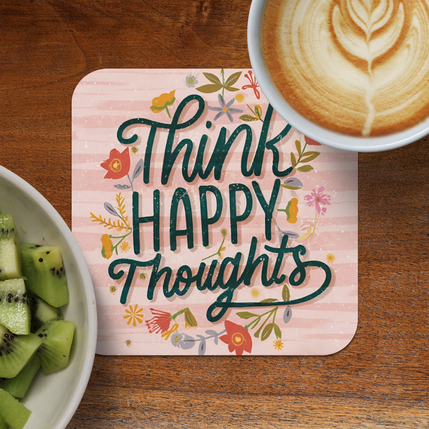 Coaster with "Think Happy Thoughts" text and floral design next to coffee and kiwi.