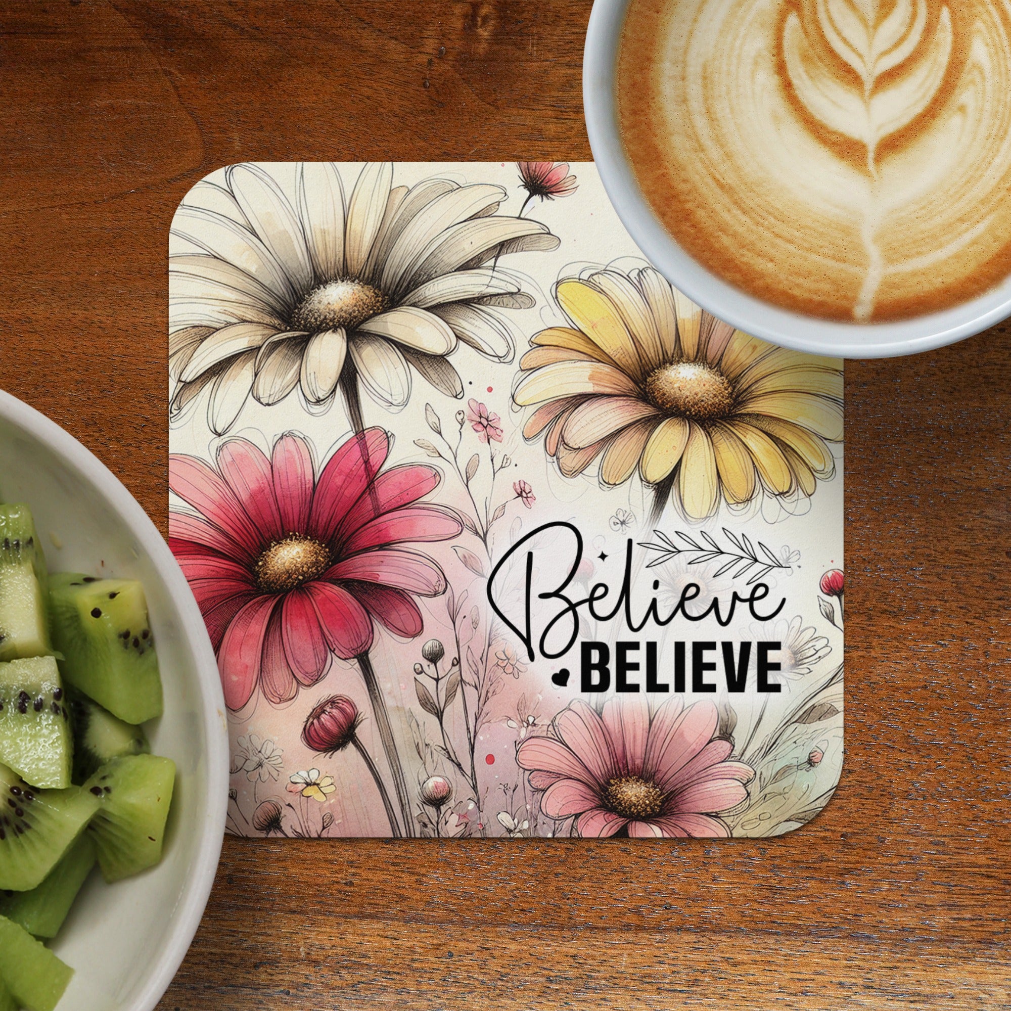 Coaster with daisy flowers and "Believe Believe" text next to kiwi slices and coffee.