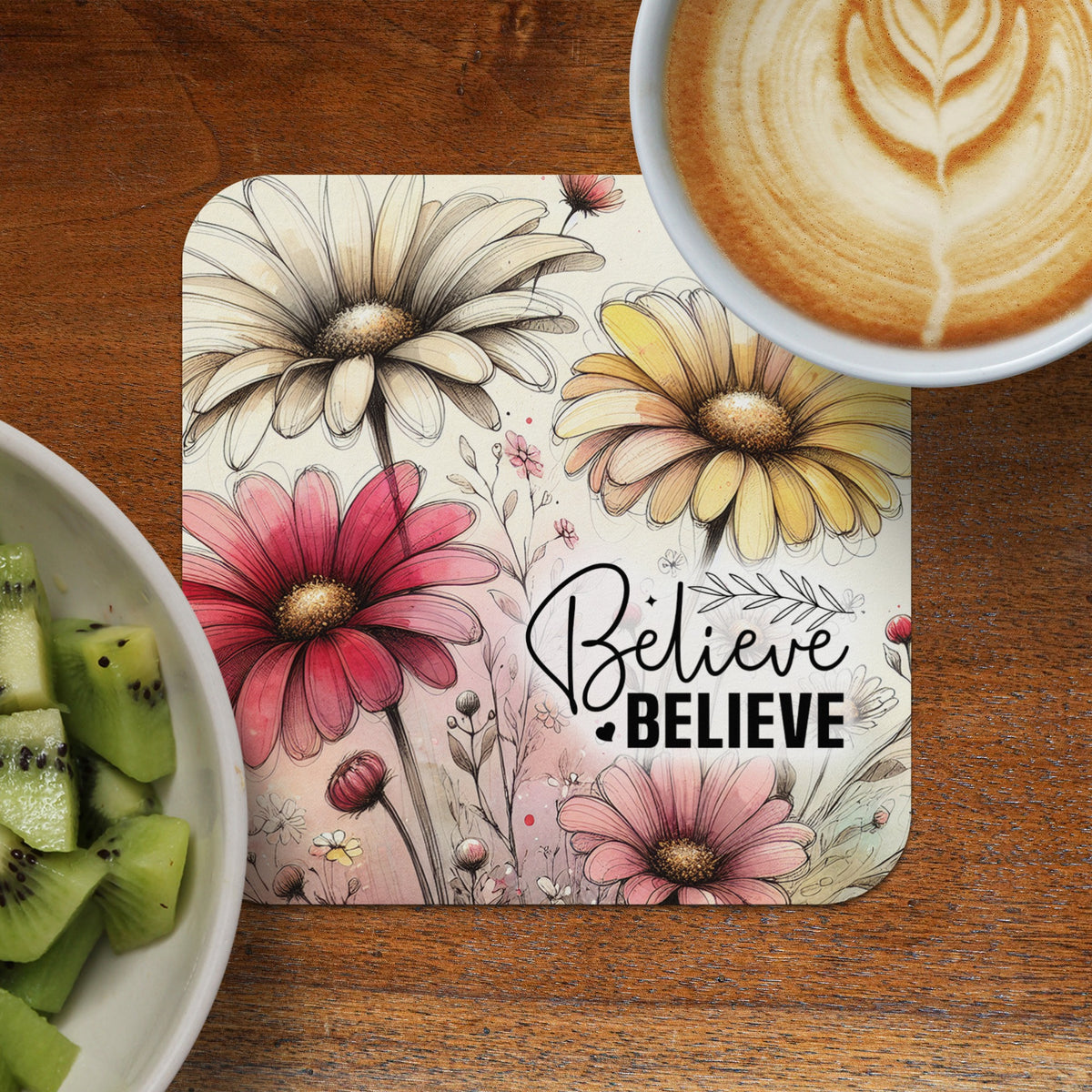 Coaster with daisy flowers and "Believe Believe" text next to kiwi slices and coffee.