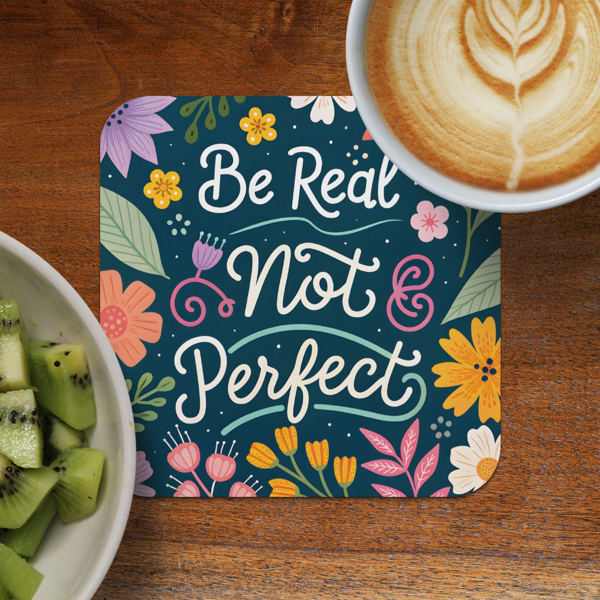 Coaster with "Be Real Not Perfect" text and floral design next to kiwi and coffee.