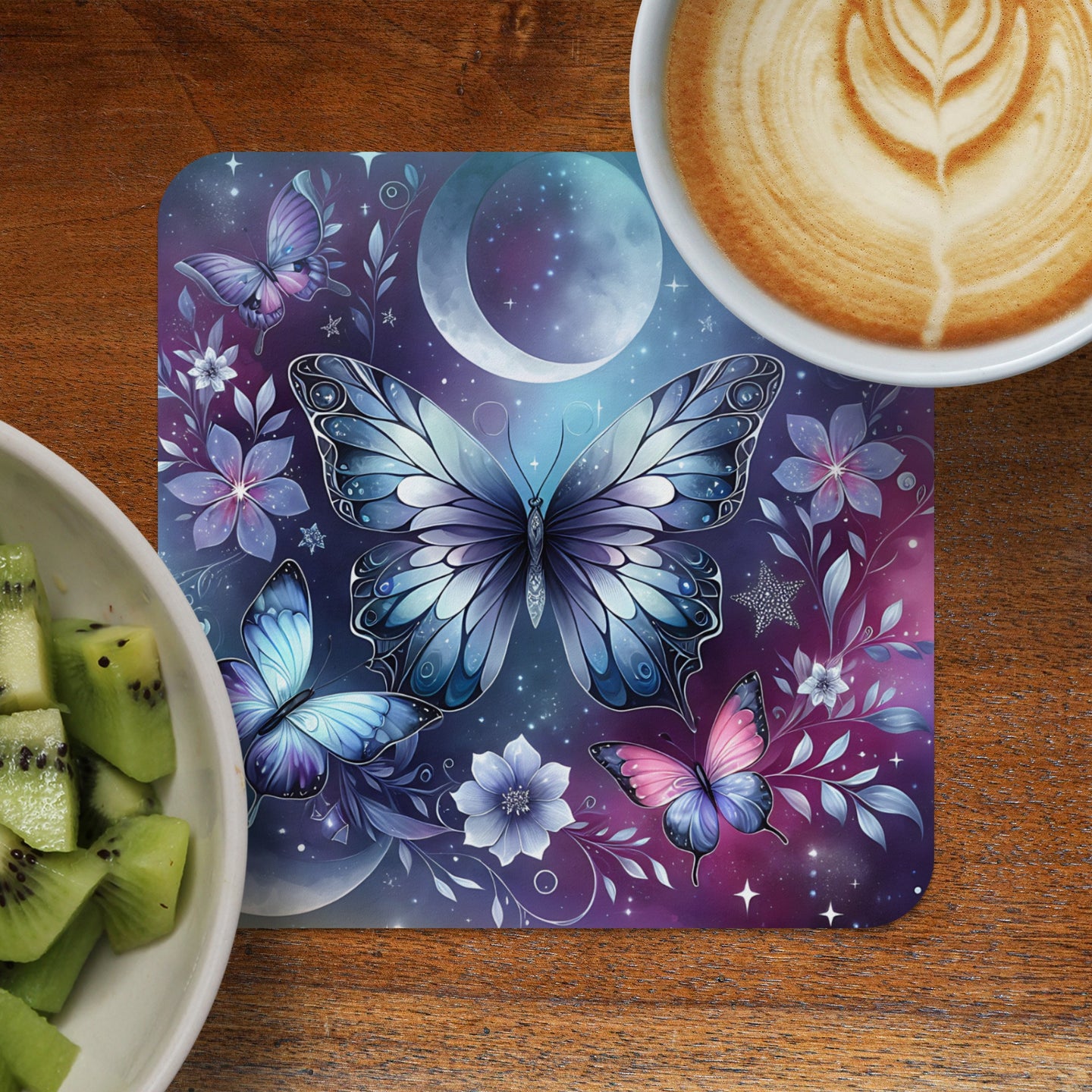 Butterfly and moon art on a coaster next to kiwi and coffee.