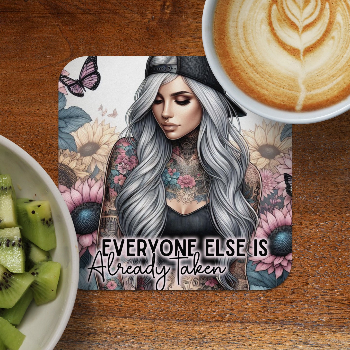 Tattooed woman with long grey hair on a coaster with floral background and text.