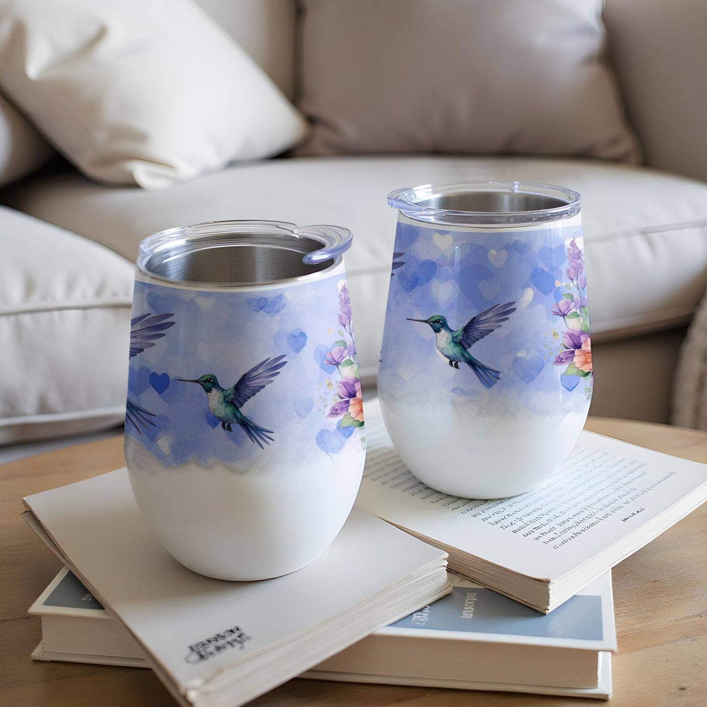 Two insulated tumblers with hummingbirds and floral patterns, resting on books.