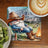 Gnome in cowboy hat and boots rests by campfire, near RV and coffee.