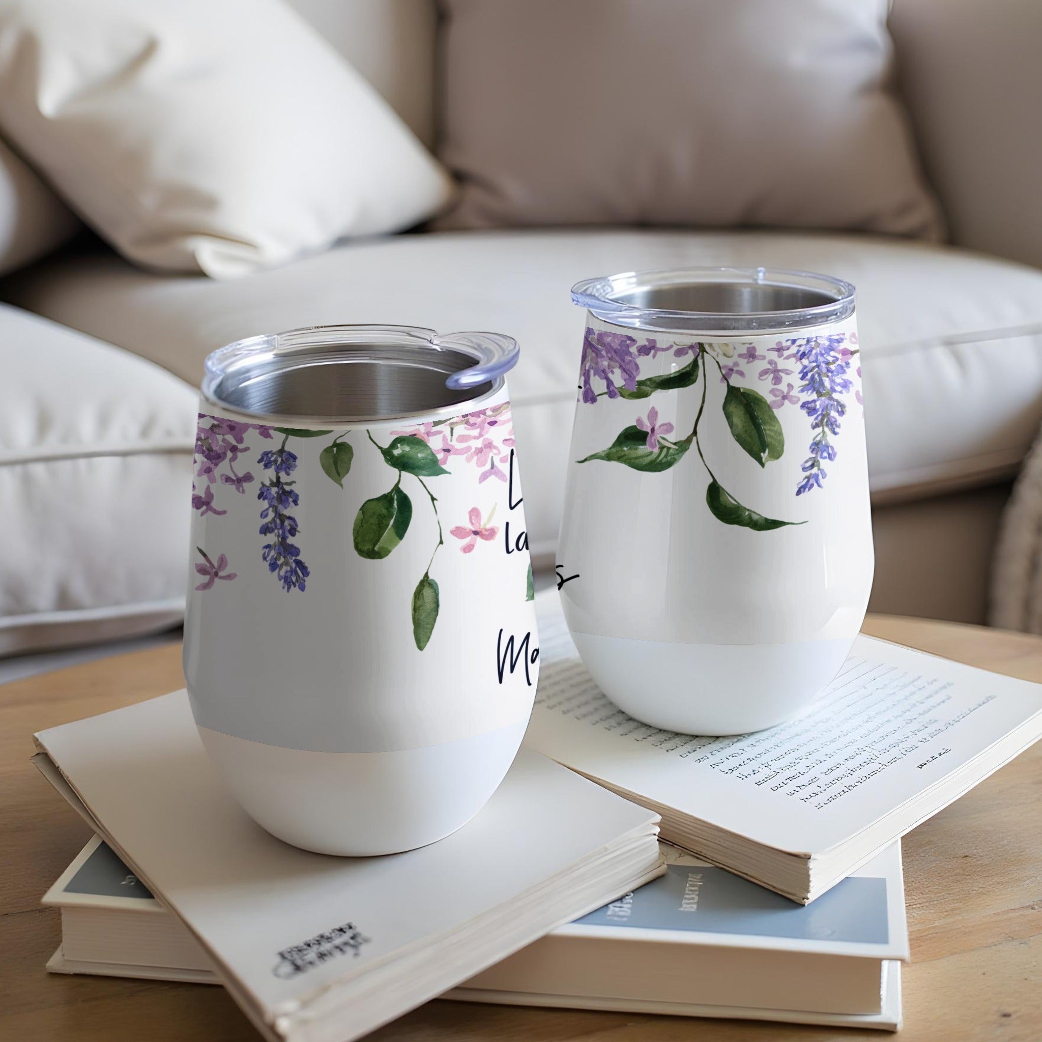 Two white tumblers with purple floral design and script text sit on books.