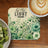 Coaster with "be the light" text and floral design, next to kiwi and coffee.