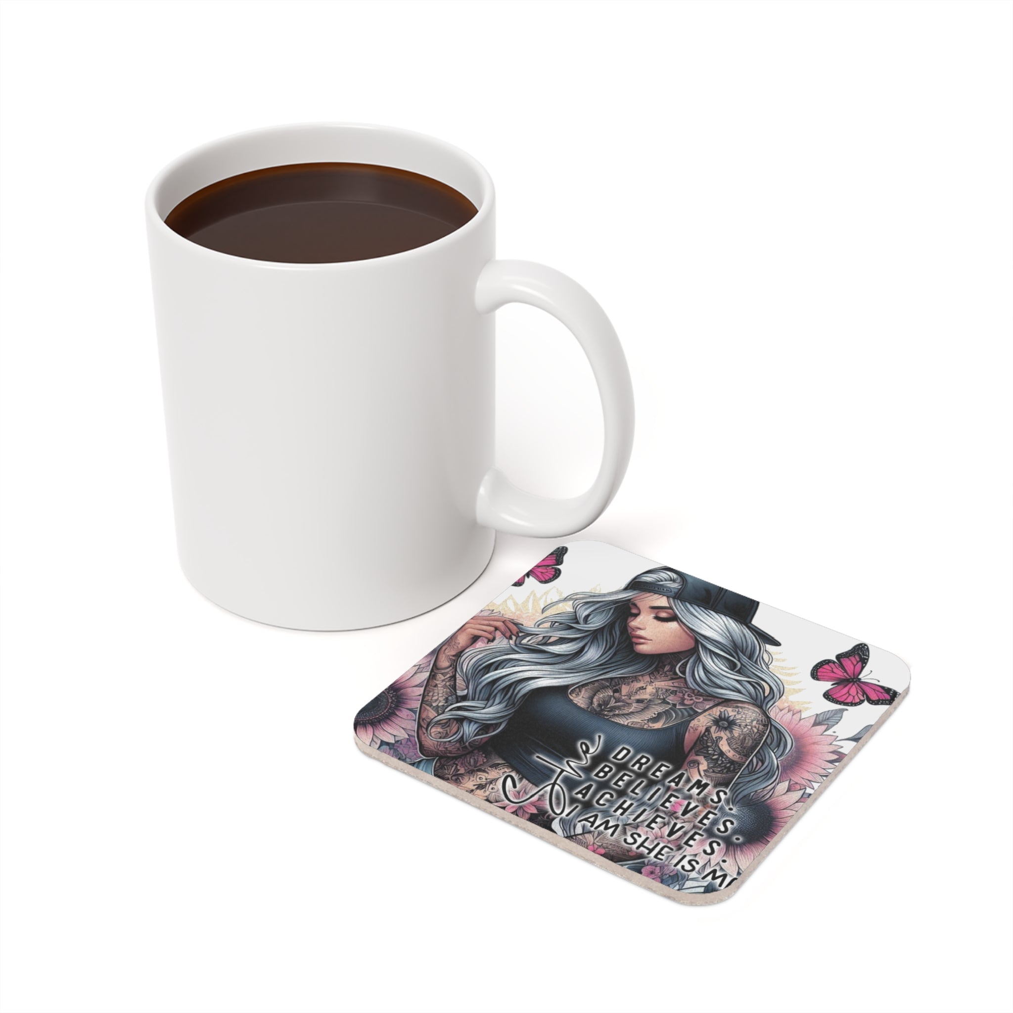 White mug of coffee next to a coaster with butterflies and text "DREAMS BELIEVE ACHIEVE AM SHE IS M".
