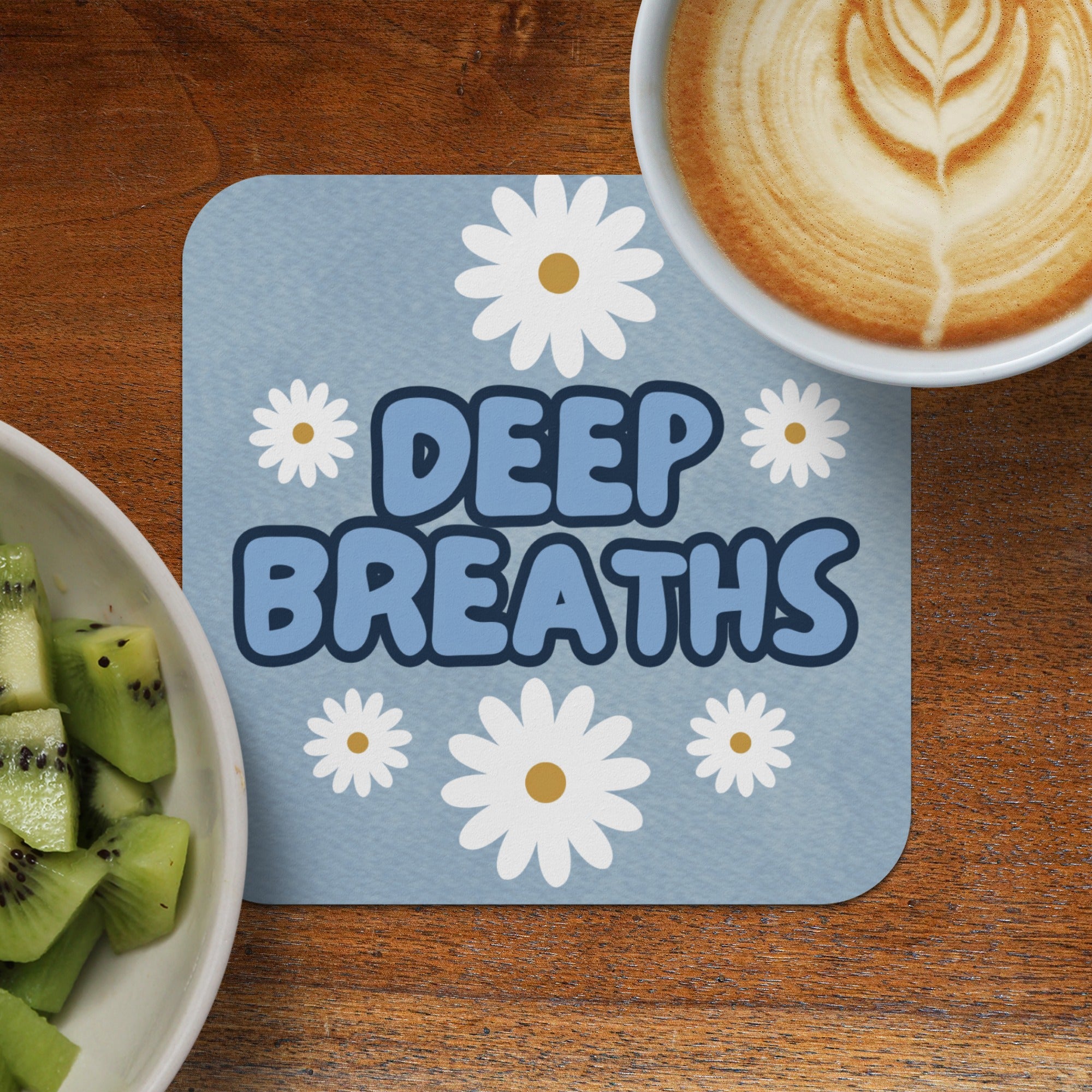 Blue coaster with "DEEP BREATHS" and daisies, next to coffee and kiwi.