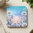 Square coaster with daisies and sky scene on a textured surface.