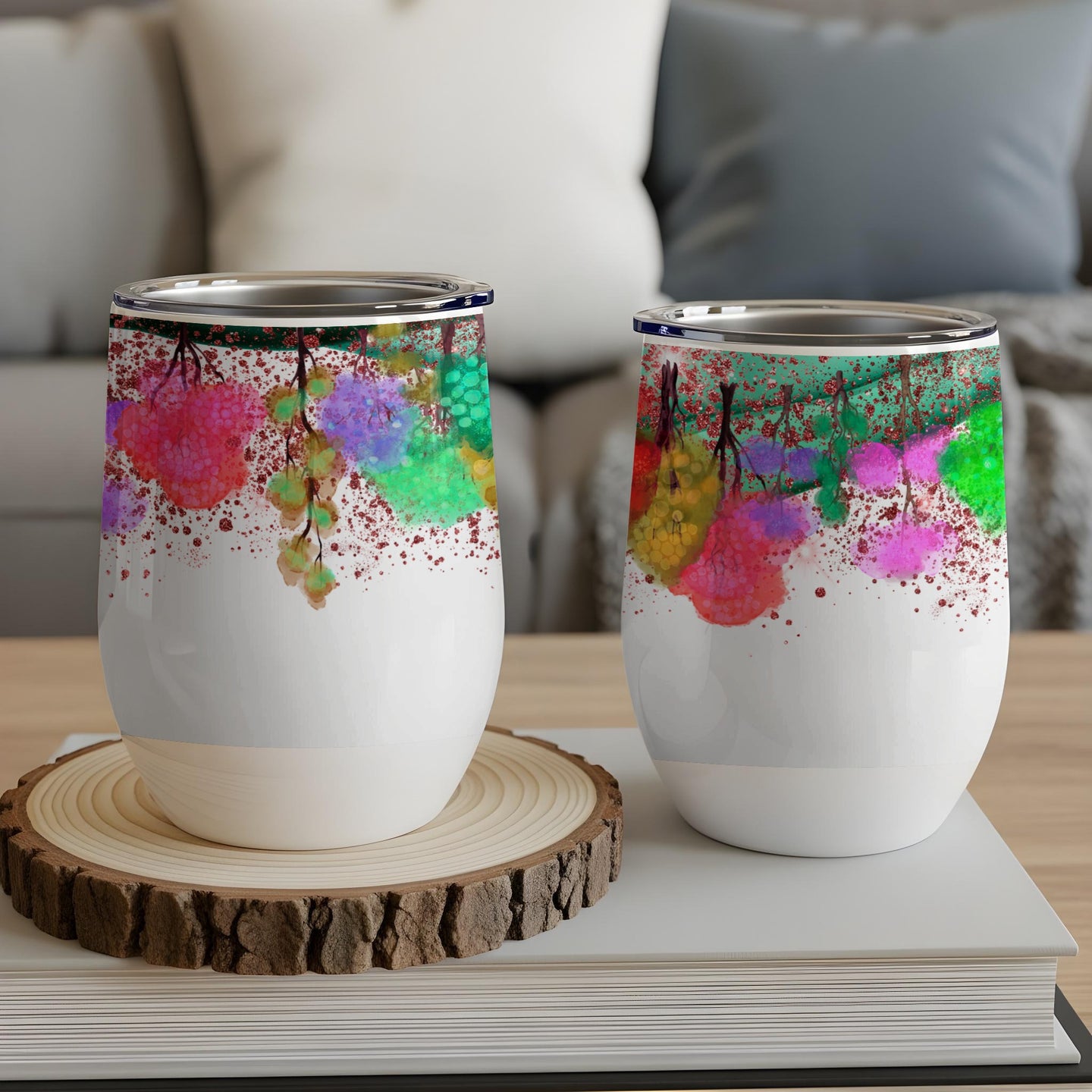 Two white tumblers with colorful abstract floral art, on a wooden coaster and books.