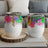 Two white tumblers with colorful abstract floral art, on a wooden coaster and books.
