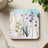 Square coaster with a watercolor floral design featuring delicate wildflowers and leaves.