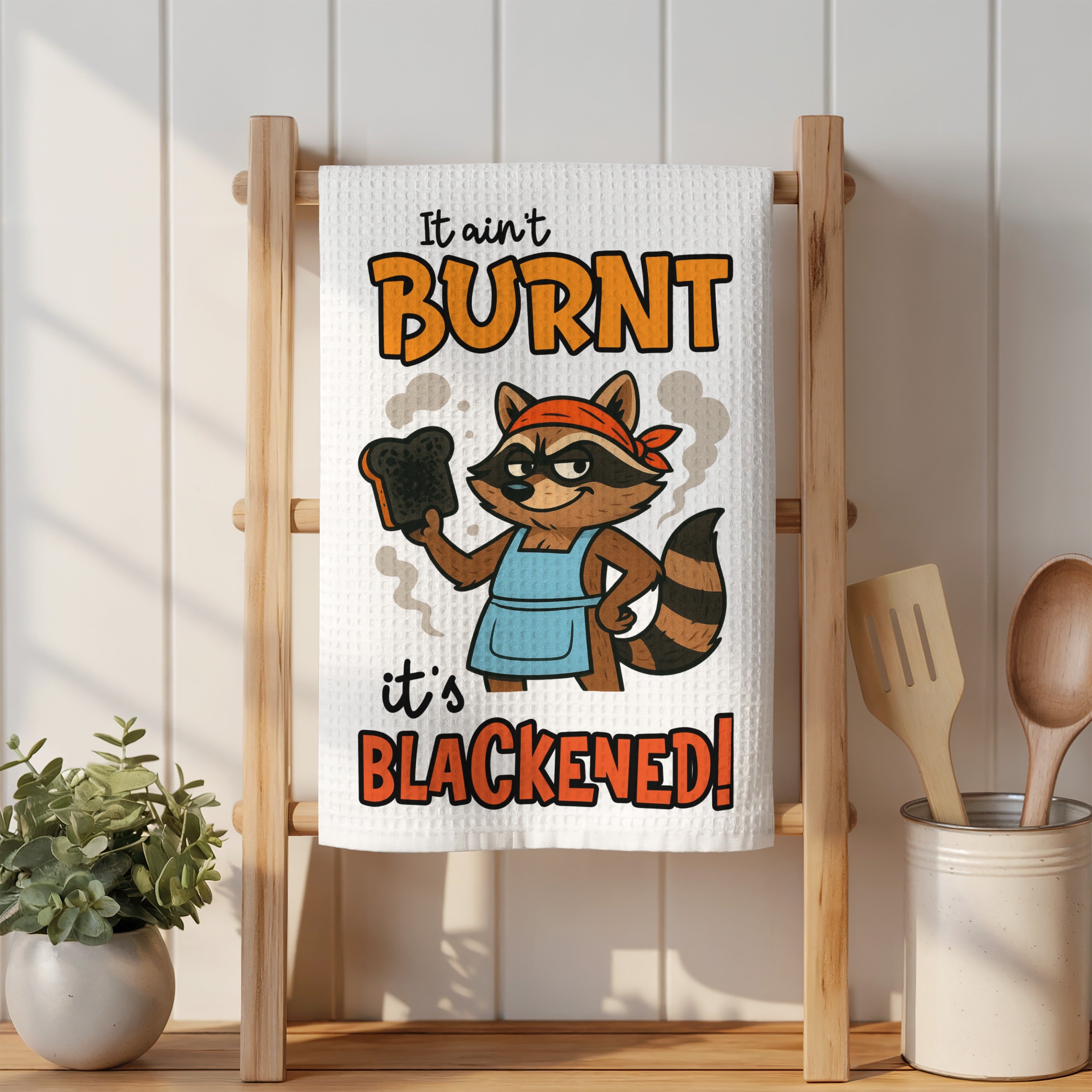 Kitchen towel with a raccoon saying "It ain't burnt, it's blackened!"