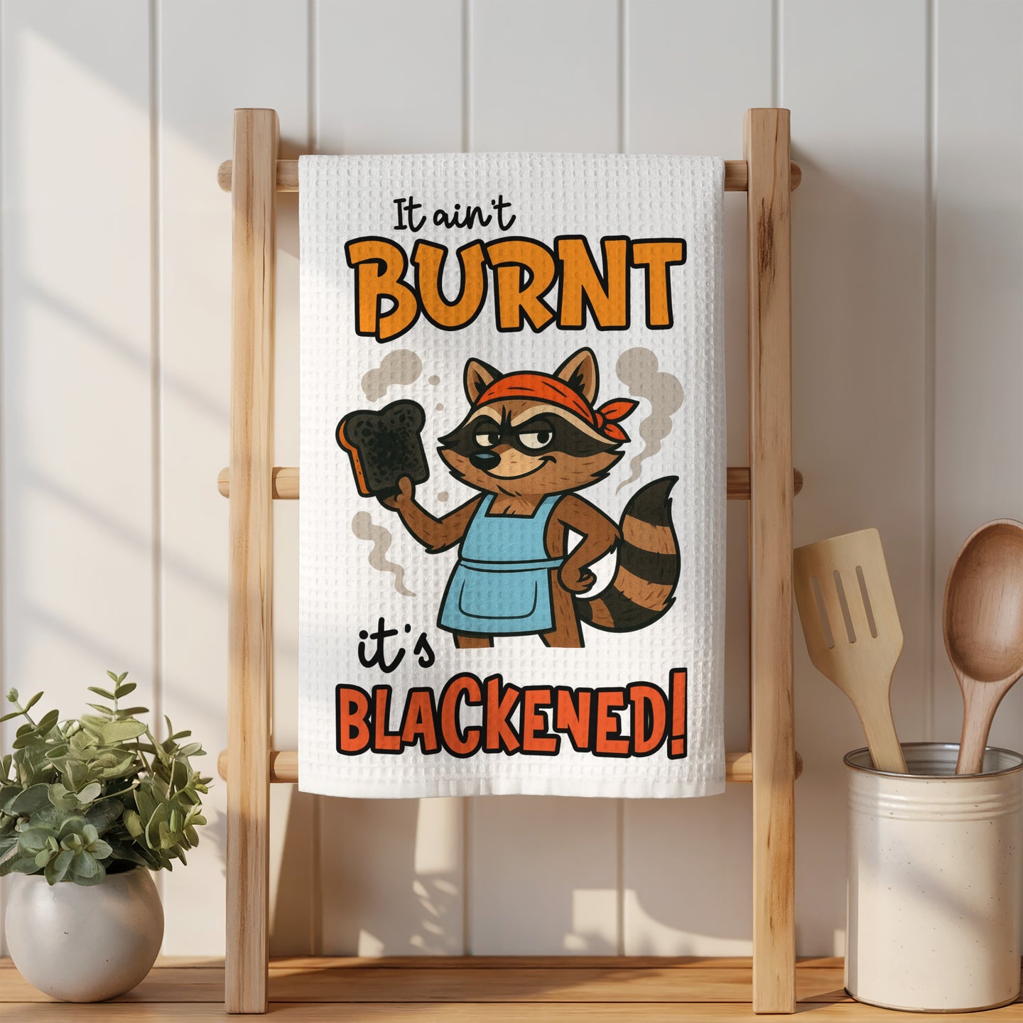 Kitchen towel with a raccoon saying "It ain't burnt, it's blackened!"