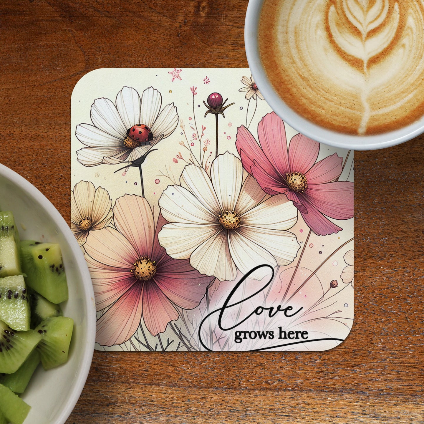 Cosmos flowers and ladybug on coaster with "love grows here" text, next to coffee and kiwi.