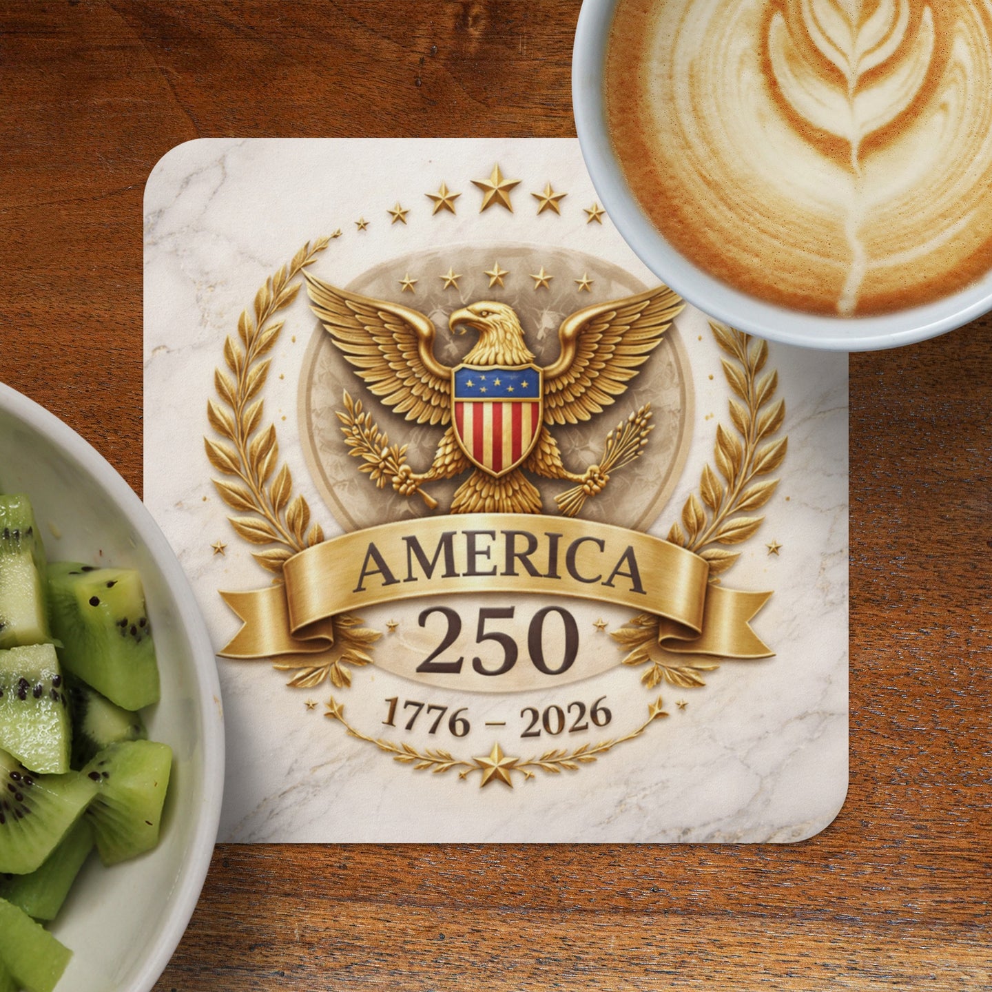 Golden eagle crest with "AMERICA 1776 - 2026" text on marble coaster, coffee, and kiwi.