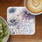 Square coaster with purple and blue floral design and text "live a great story".