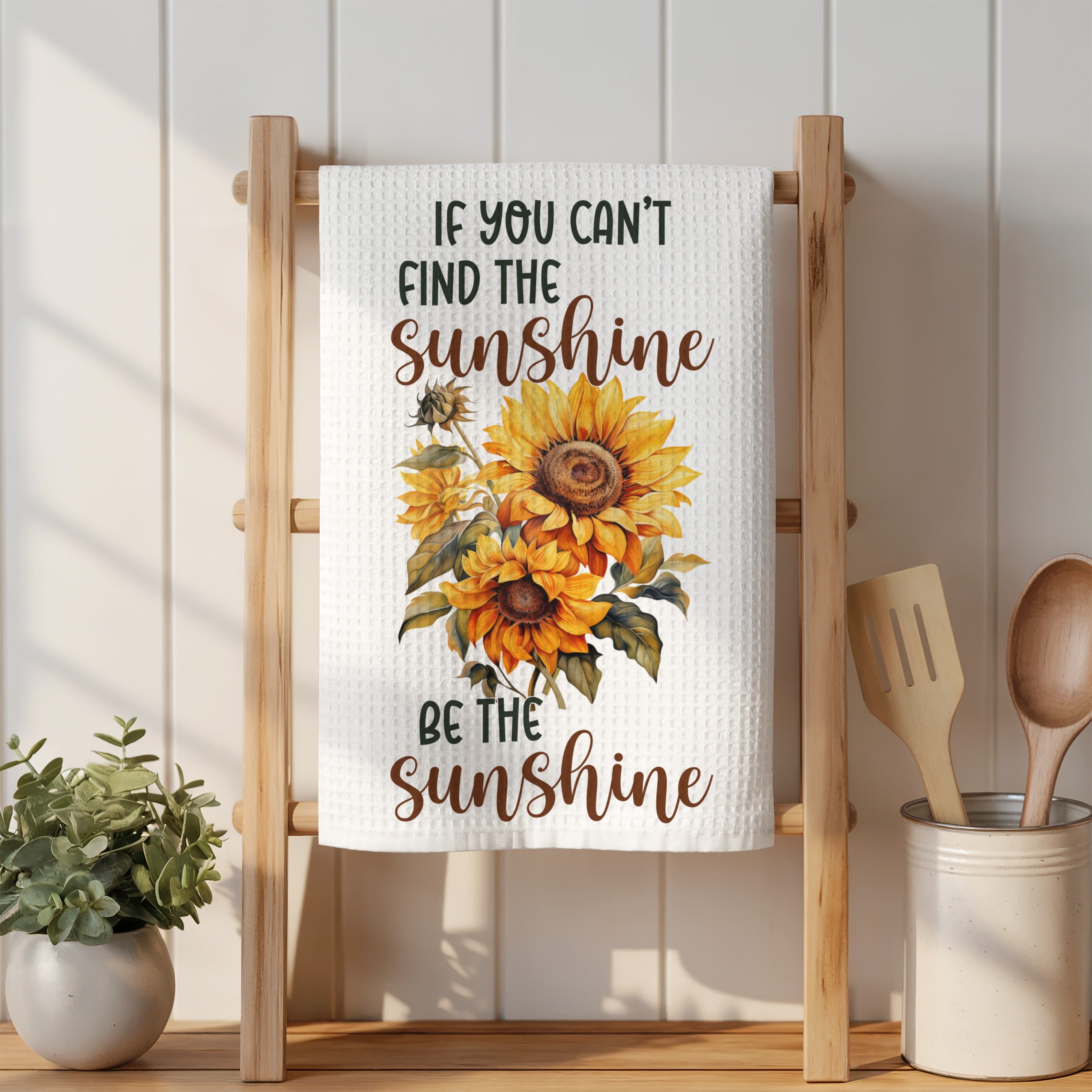 Sunflower art and quote "If you can't find the sunshine, be the sunshine" on a kitchen towel.