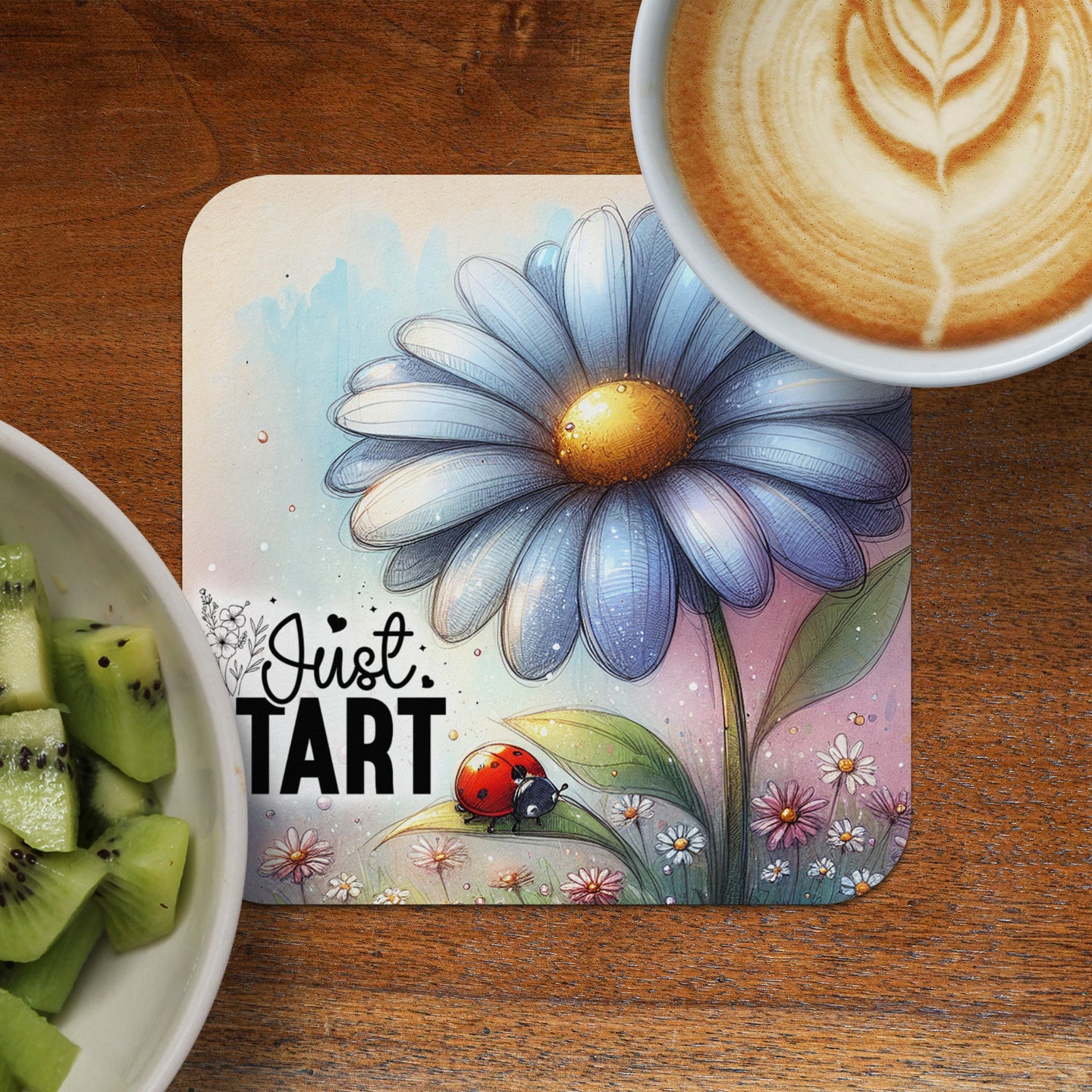 Coaster with blue daisy, ladybug, and text "Just Tart" next to a coffee and kiwi slices.