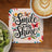 Square coaster with "Smile and Shine" text, butterflies, and flowers, next to kiwi and coffee.