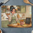 Woman in apron holding spoons in kitchen with fried chicken and chalk sign.