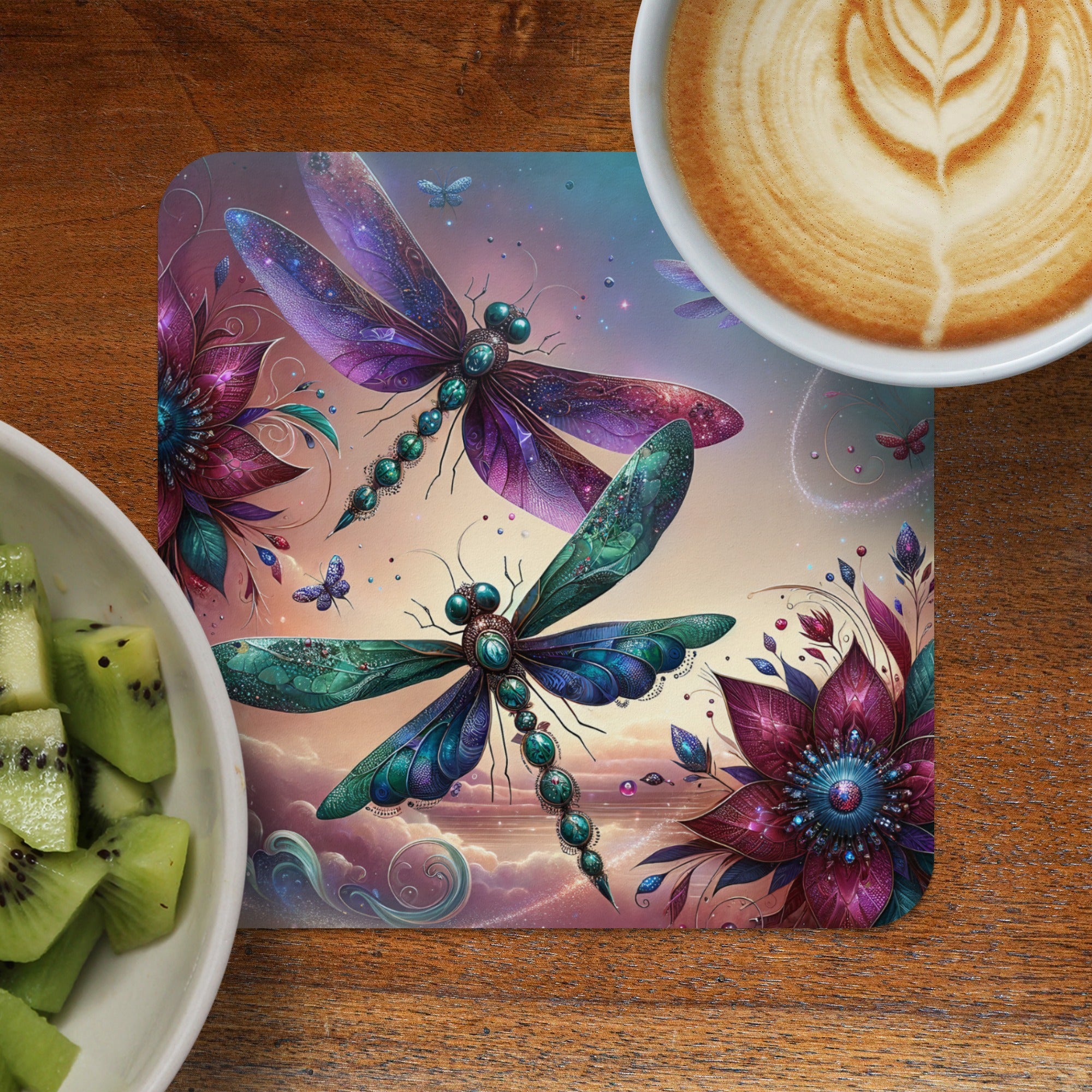 Dragonflies and ornate flowers on a square coaster with a latte and kiwi slices.