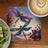Dragonflies and ornate flowers on a square coaster with a latte and kiwi slices.