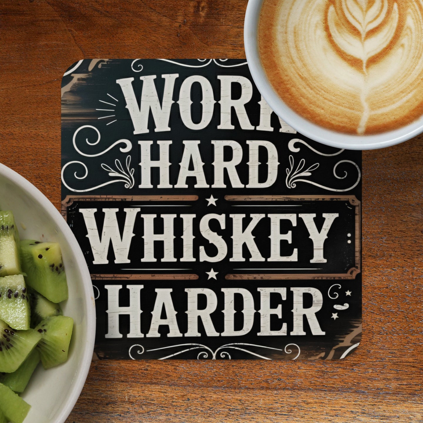 Wooden coaster with "WORK HARD WHISKEY HARDER" text next to kiwi slices and coffee.