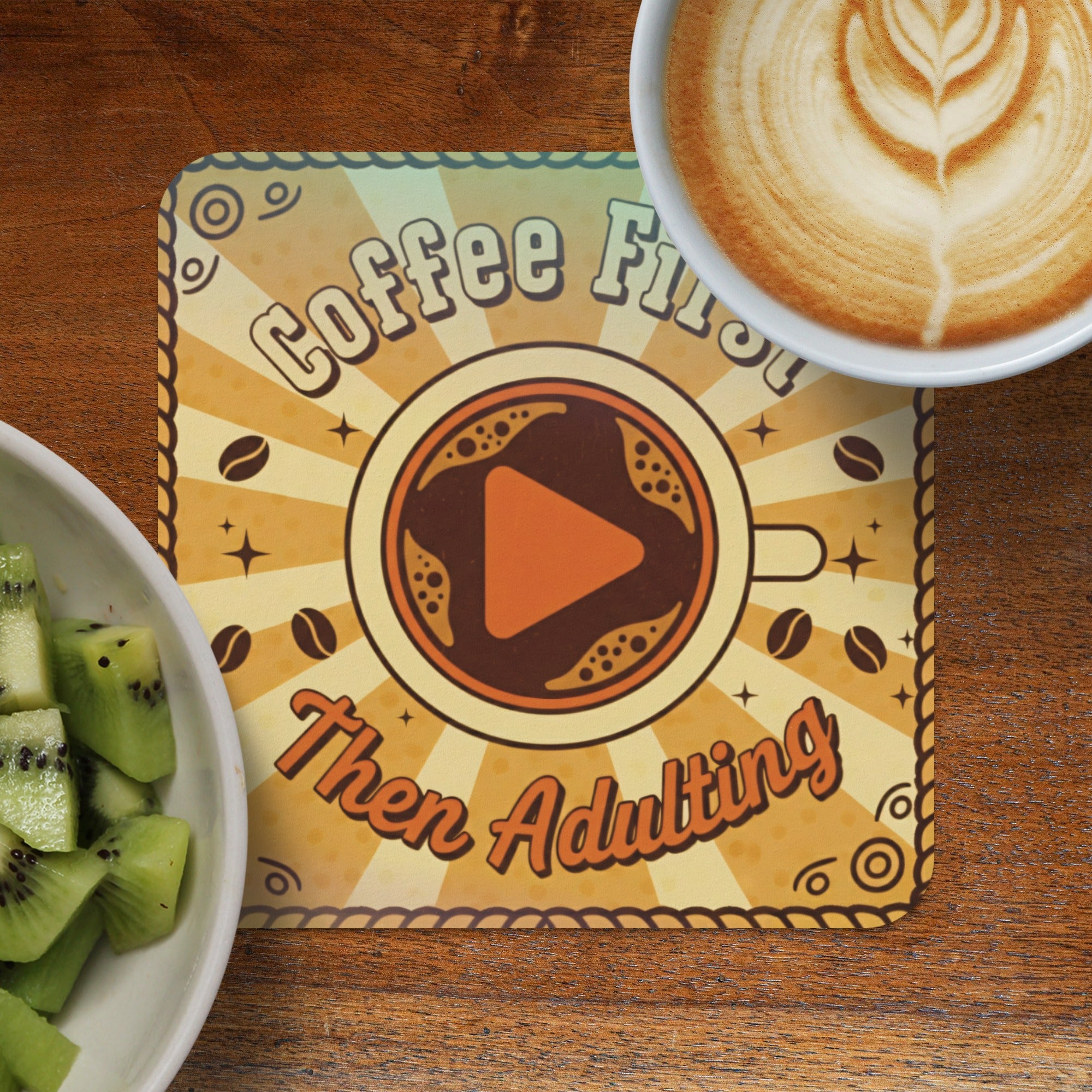 Coffee First Then Adulting coaster with play button, next to kiwi fruit and coffee.