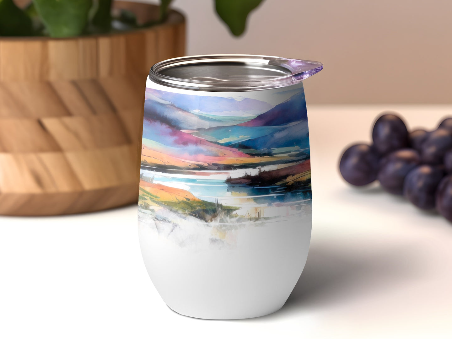 White tumbler with colorful landscape art, purple grapes and plant in background.