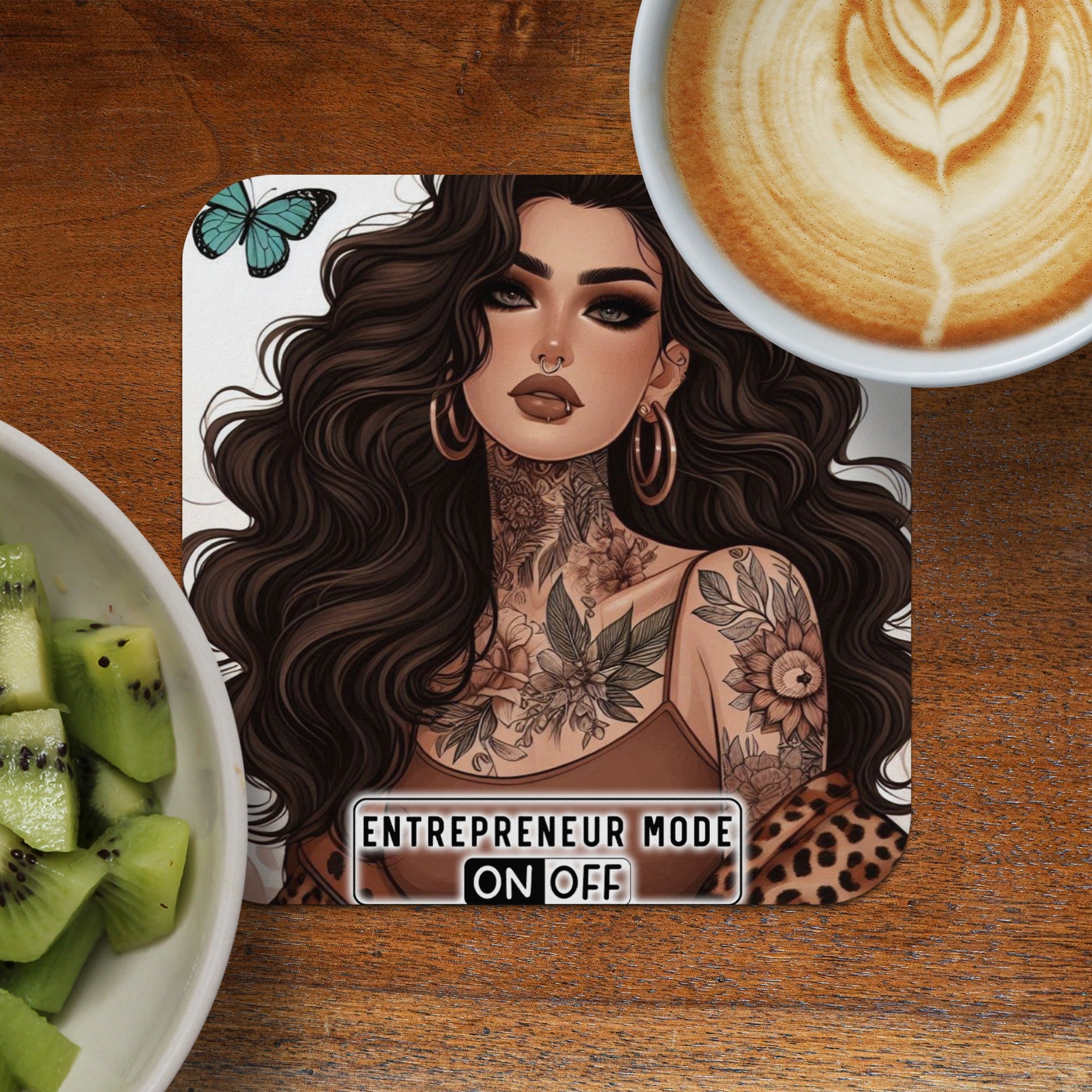 Woman with tattoos and "Entrepreneur Mode On Off" text on a coaster near kiwi and coffee.