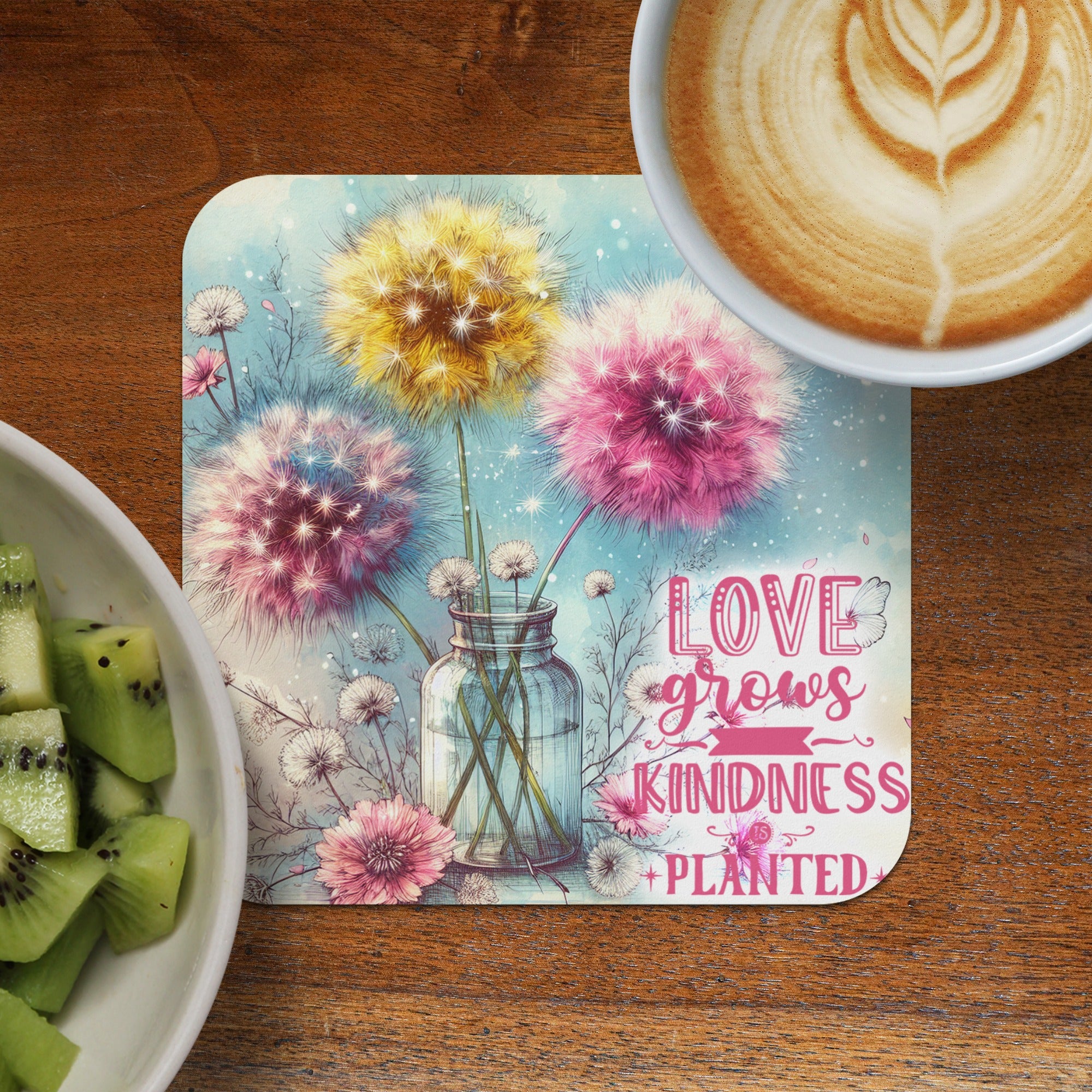 Dandelion art with inspirational text "Love grows kindness" on a coaster next to coffee and kiwi.