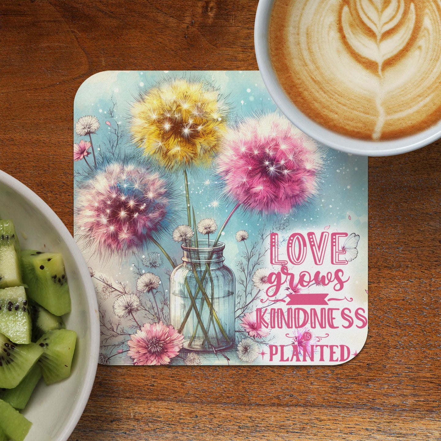 Dandelion art with inspirational text "Love grows kindness" on a coaster next to coffee and kiwi.